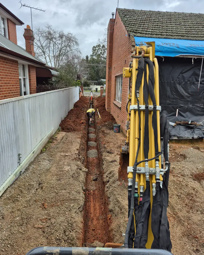 Excavator digging a sewer drain trench beside a brick home